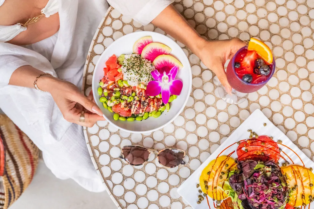 A table setting with a salad bowl, tropical fruit slices, a berry drink, and colorful plated food laid out on a patterned surface.