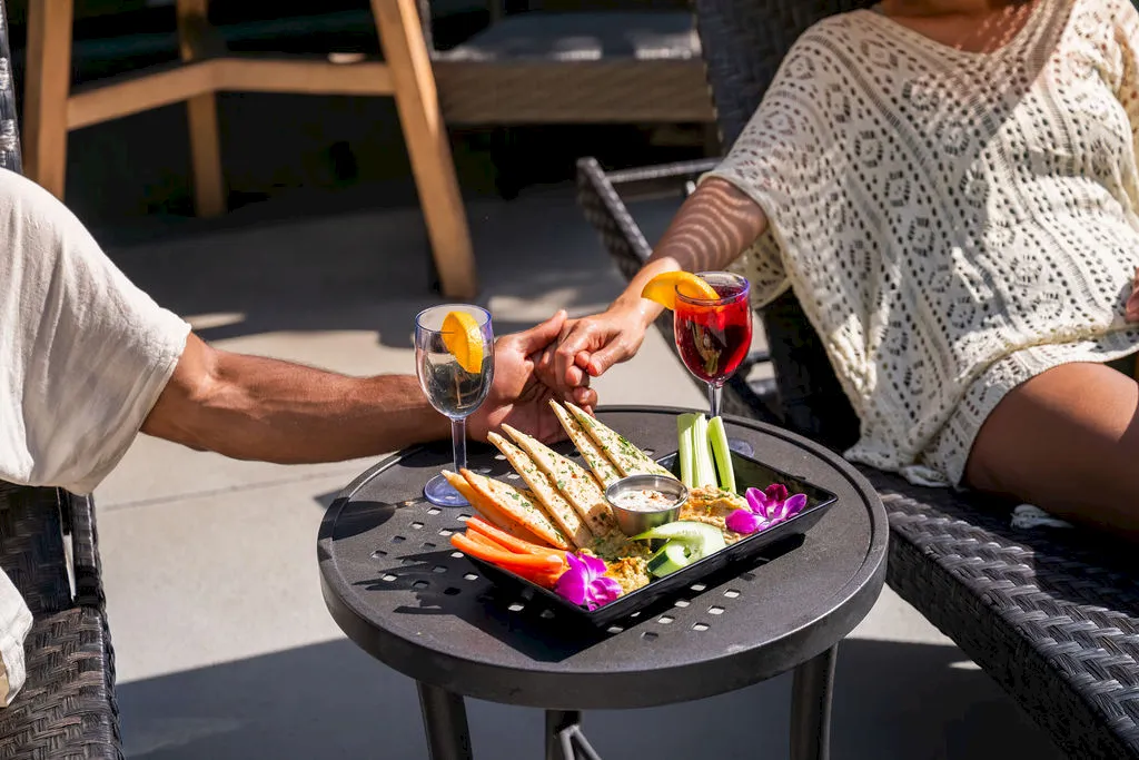 Two people clink glasses over a grill plate with colorful vegetables and purple flowers at an outdoor dining table.
