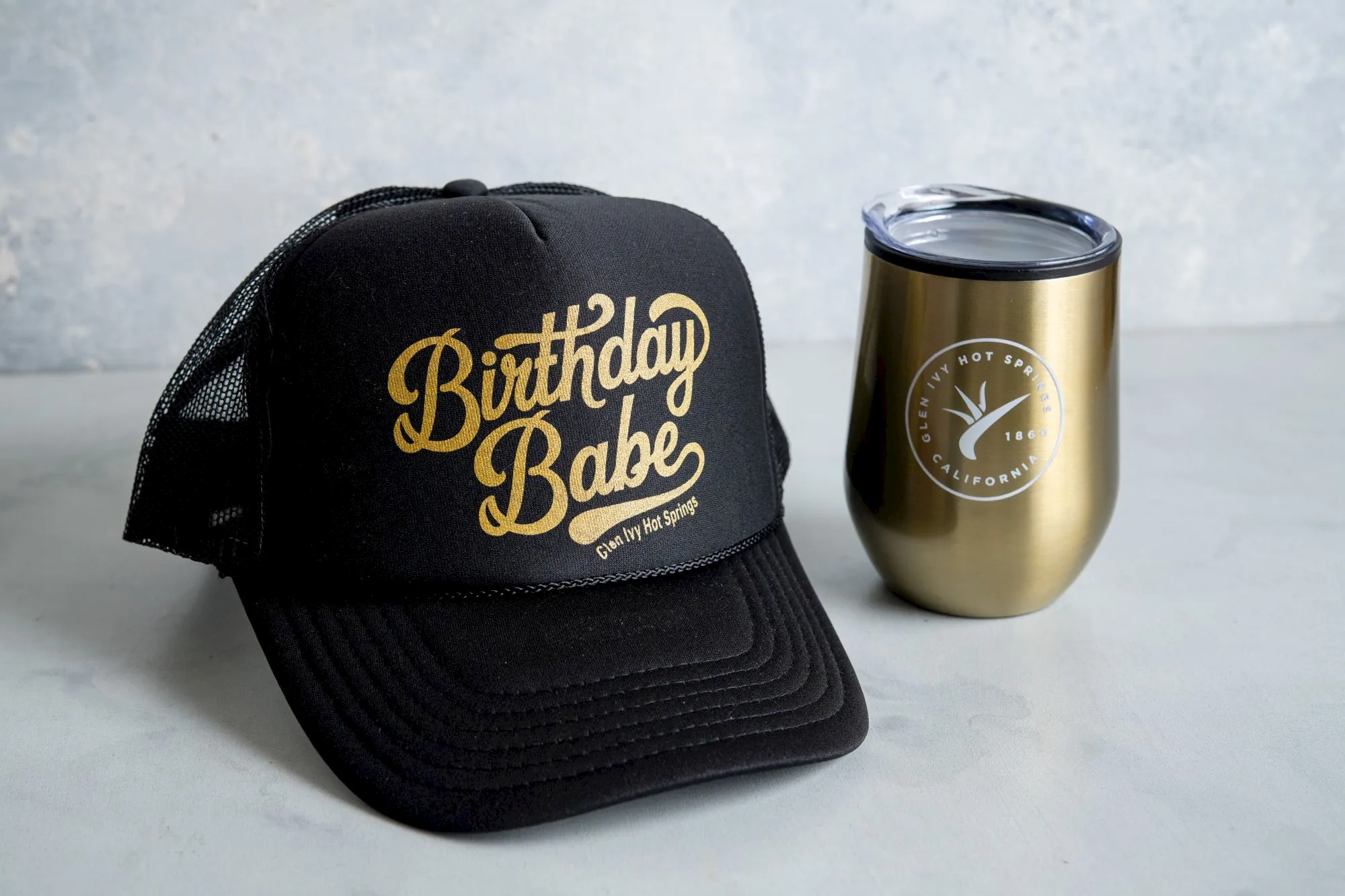 A black &ldquo;Birthday Babe&rdquo; cap sits beside a gold &ldquo;Hot Springs 1860&rdquo; tumbler on a light background, both branded for hot sauce.