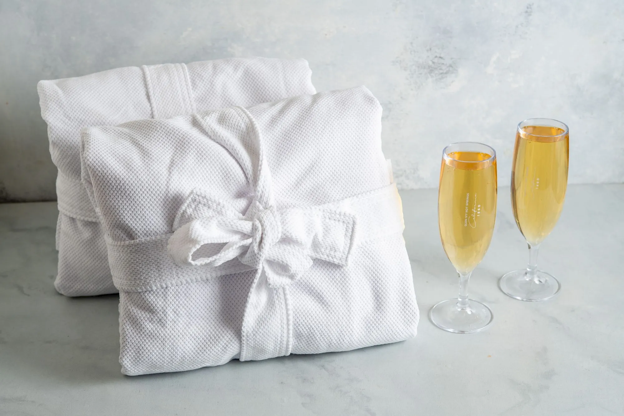 A white spa robe is folded beside two champagne flutes filled with golden sparkling wine, on a light gray surface.