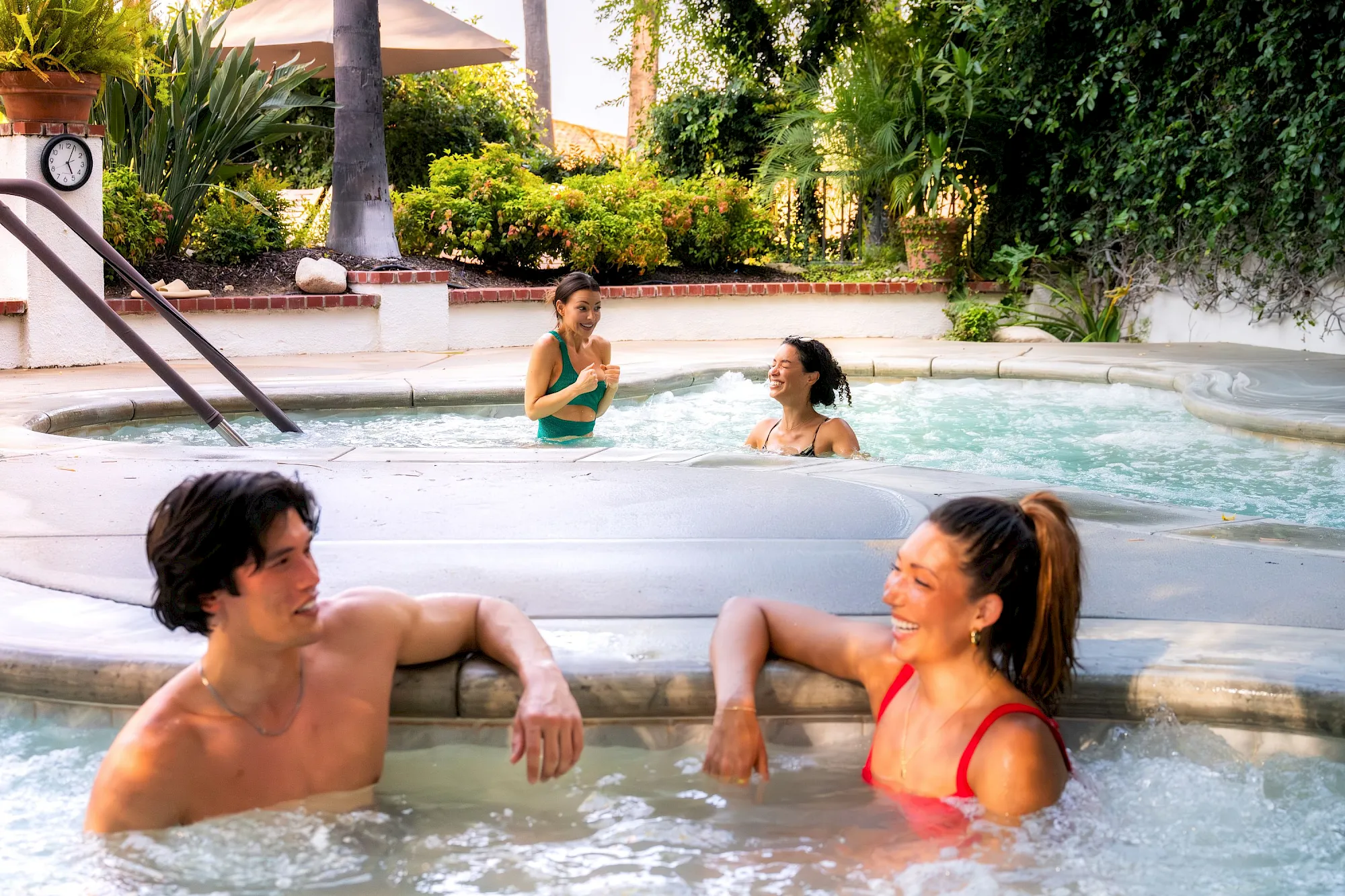 Friends relax and laugh in a hot tub surrounded by lush tropical gardens under warm sunlight.