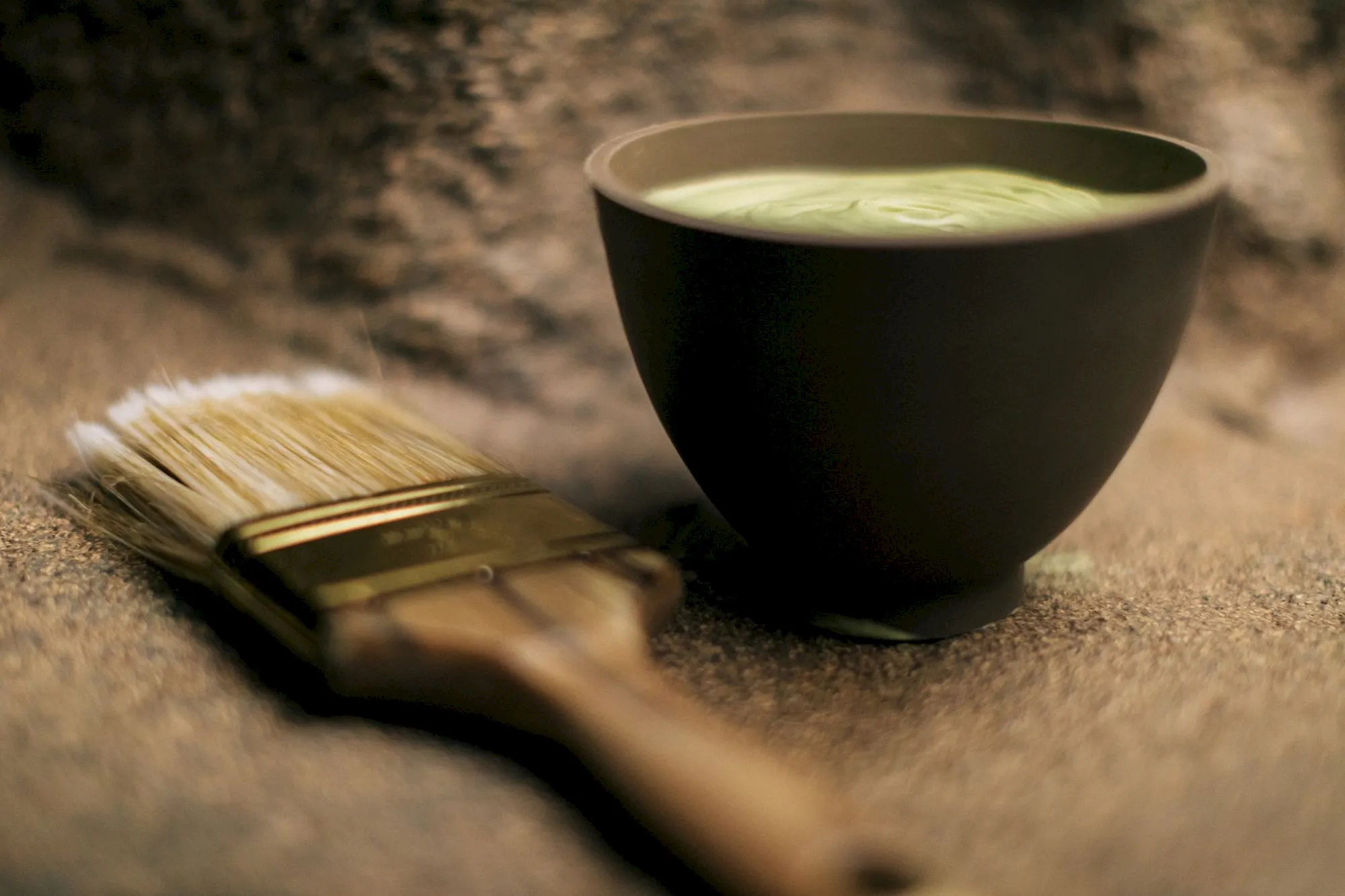 A dark ceramic bowl filled with matcha-green tea sits on a stone surface beside a bamboo whisk.