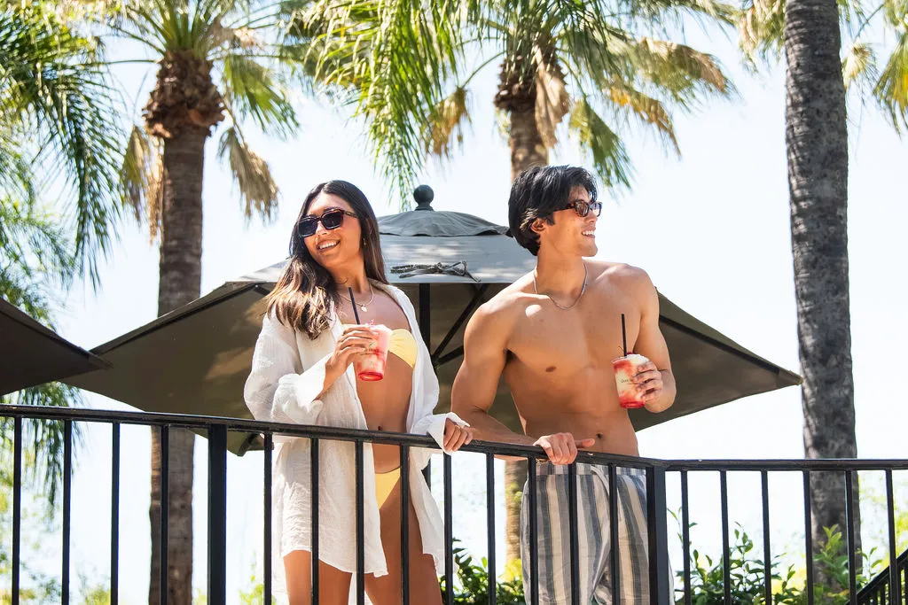Two friends relax on a sunny beach deck with umbrellas, smiling and sipping colorful drinks in the shade, basking in the tropical vibe.