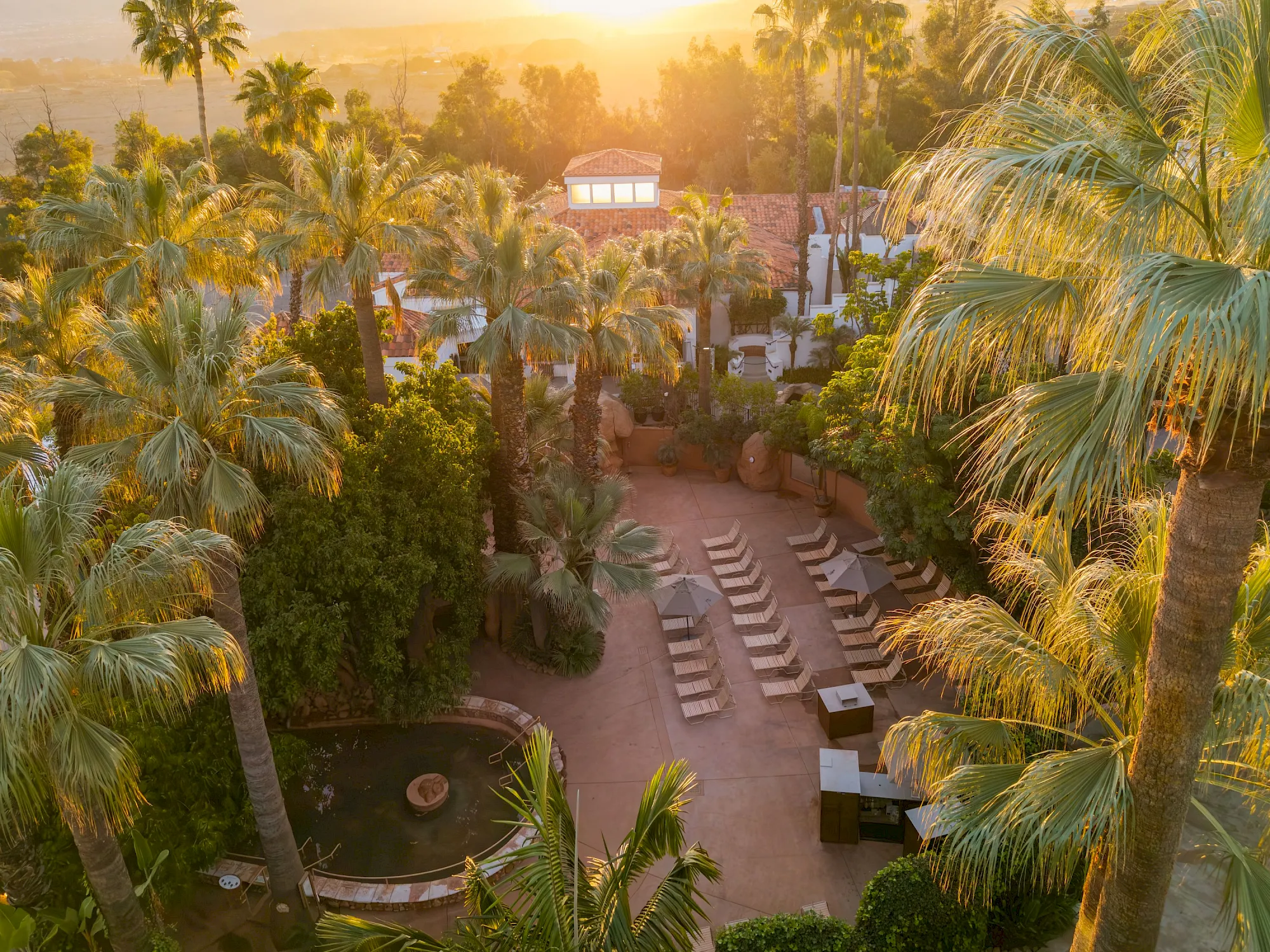 A tropical resort at golden sunrise with palm trees, a tiled courtyard, and lounge chairs by a small pool.