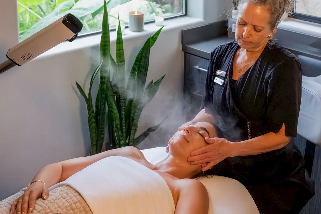 A woman performs a facial treatment on a reclining client in a spa room, using gentle hands while mist rises from the treatment area, relaxing.