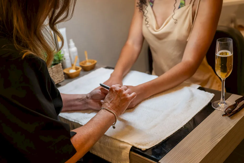 A spa massage session is underway, with a therapist gently stretching a client&rsquo;s hands while a champagne flute sits nearby, smiling.