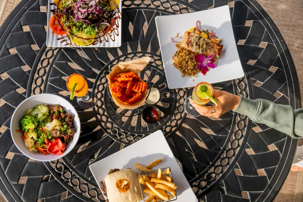 A bird&rsquo;s-eye view of four plates of colorful restaurant food&mdash;salads, pasta, a burger, fries&mdash;on a patterned metal table, with a drink, and someone&rsquo;s hand grabbing a plate.