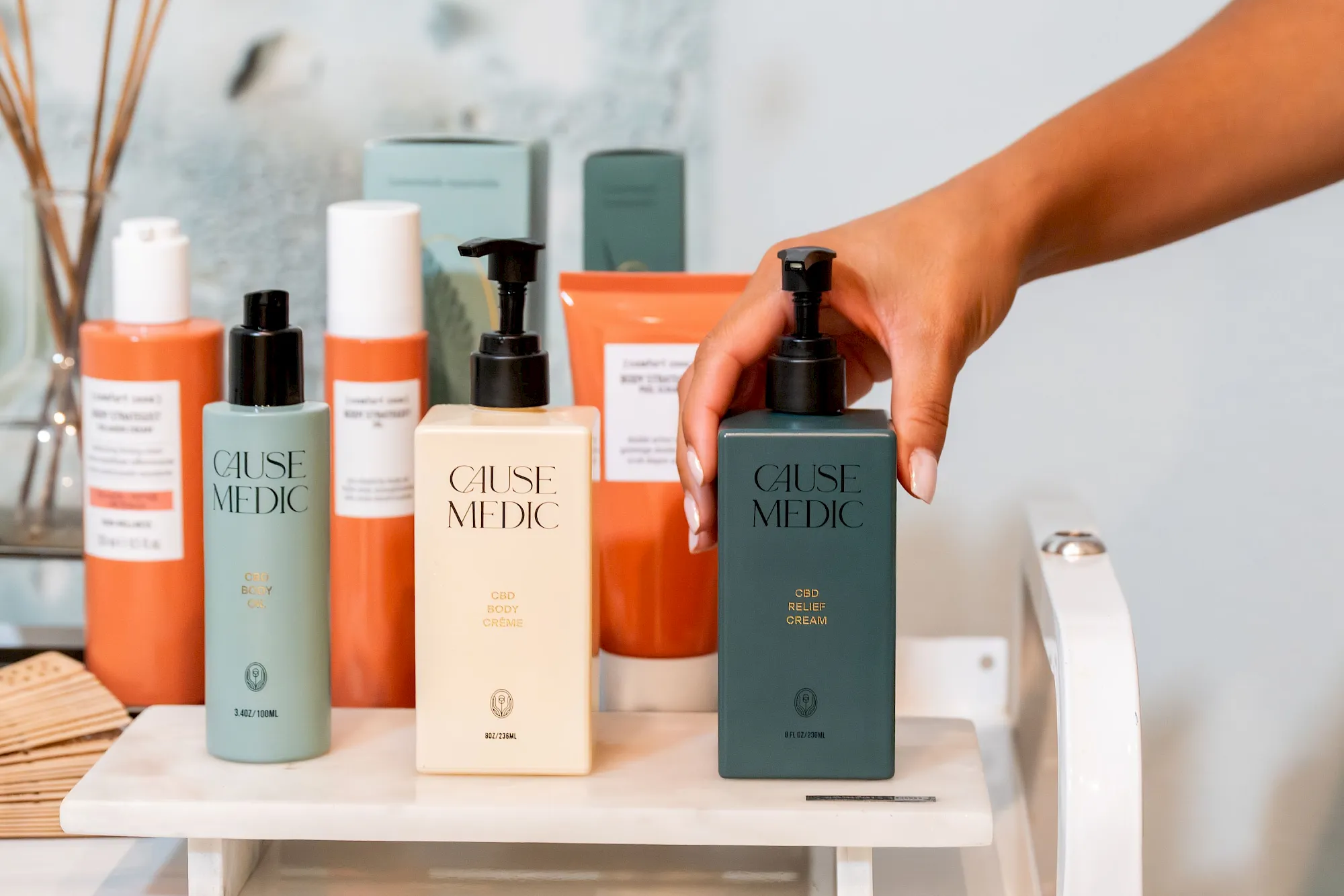 A hand holding a Cause Medic pump bottle among CBD skincare products on a bathroom shelf.
