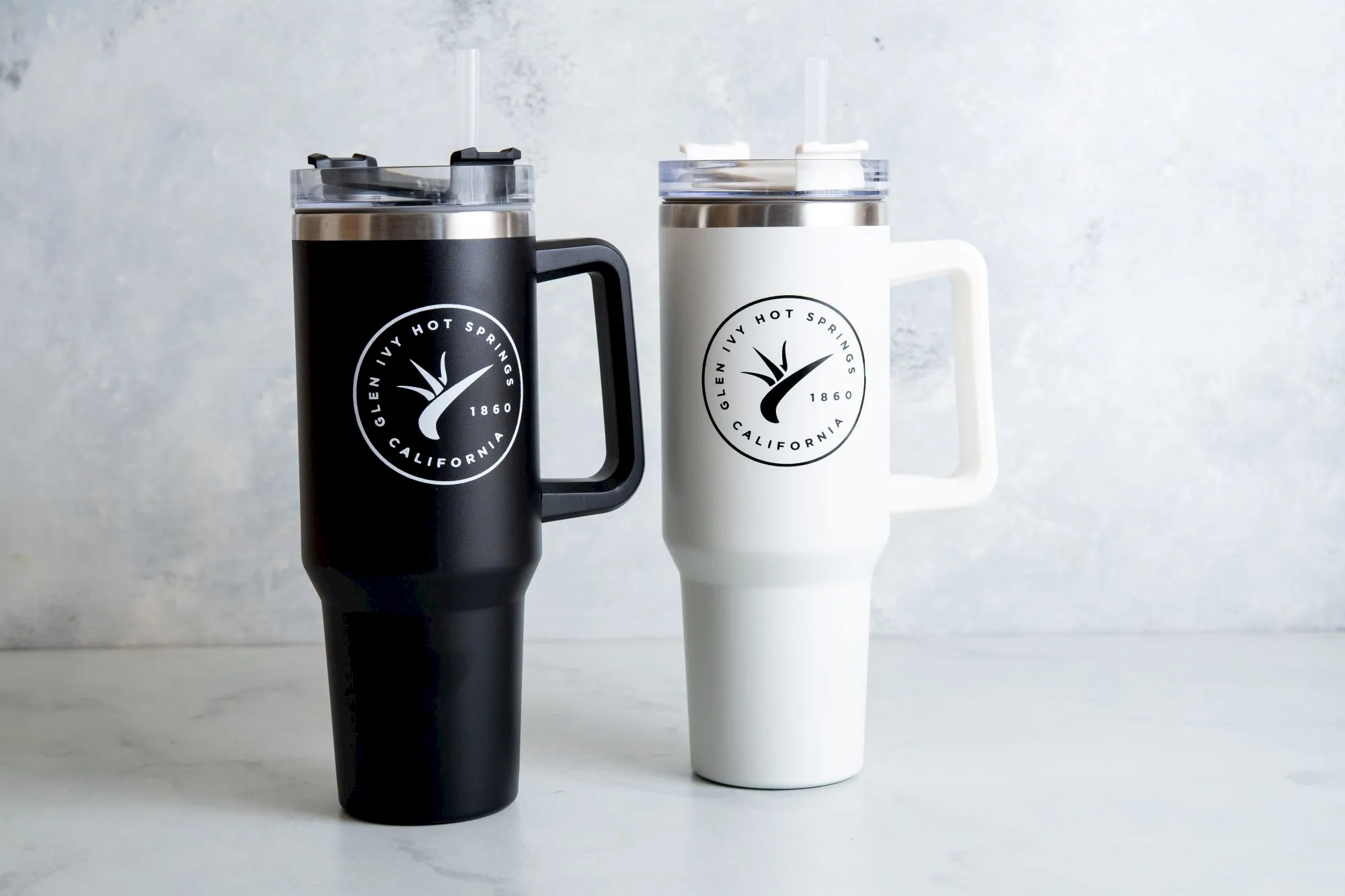 Two branded insulated travel tumblers&mdash;black and white&mdash;stand side by side with lids and straws on a light background.