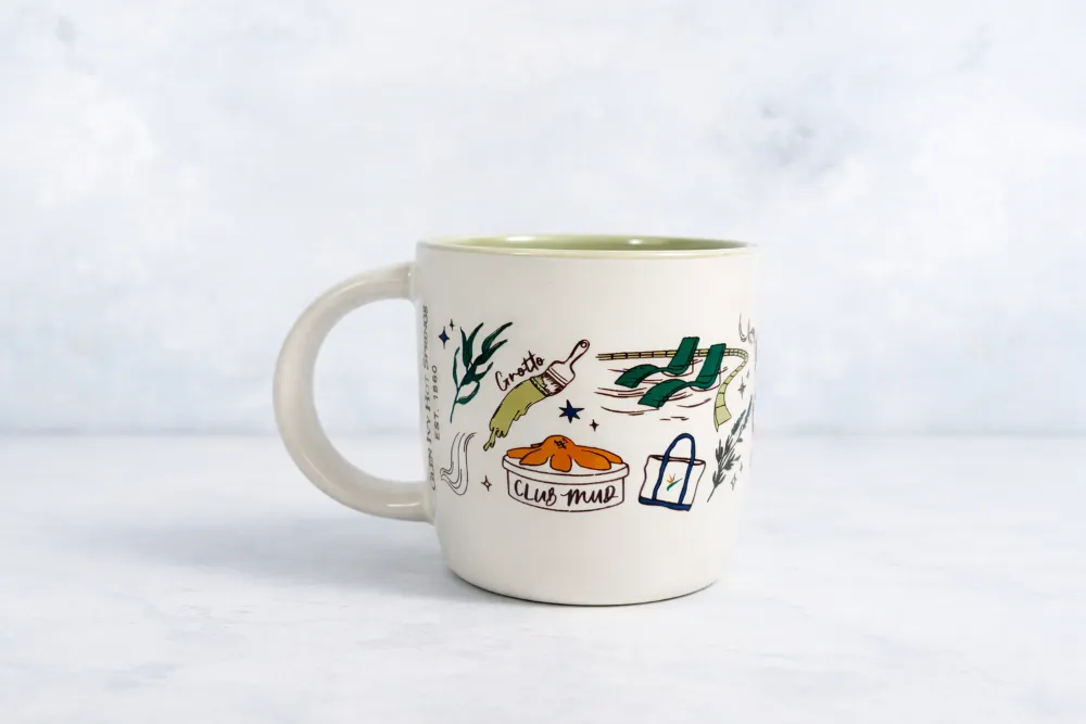 A ceramic mug on a white surface, decorated with a &ldquo;Club Mud&rdquo; design, a guitar, and green elements against a cloudy background.