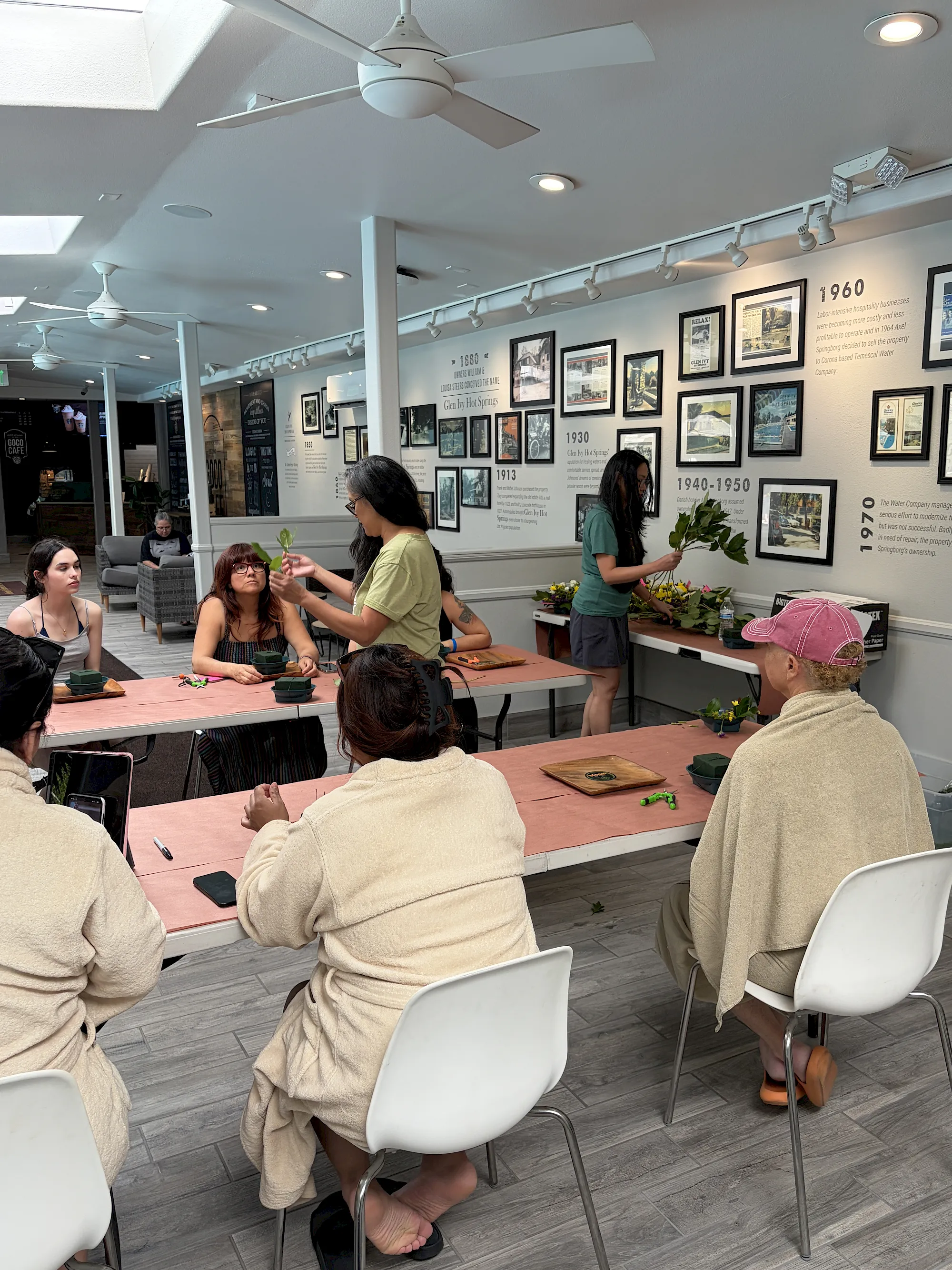 A guided activity in a museum-style room where participants sit at tables as someone presents small plants and tools.