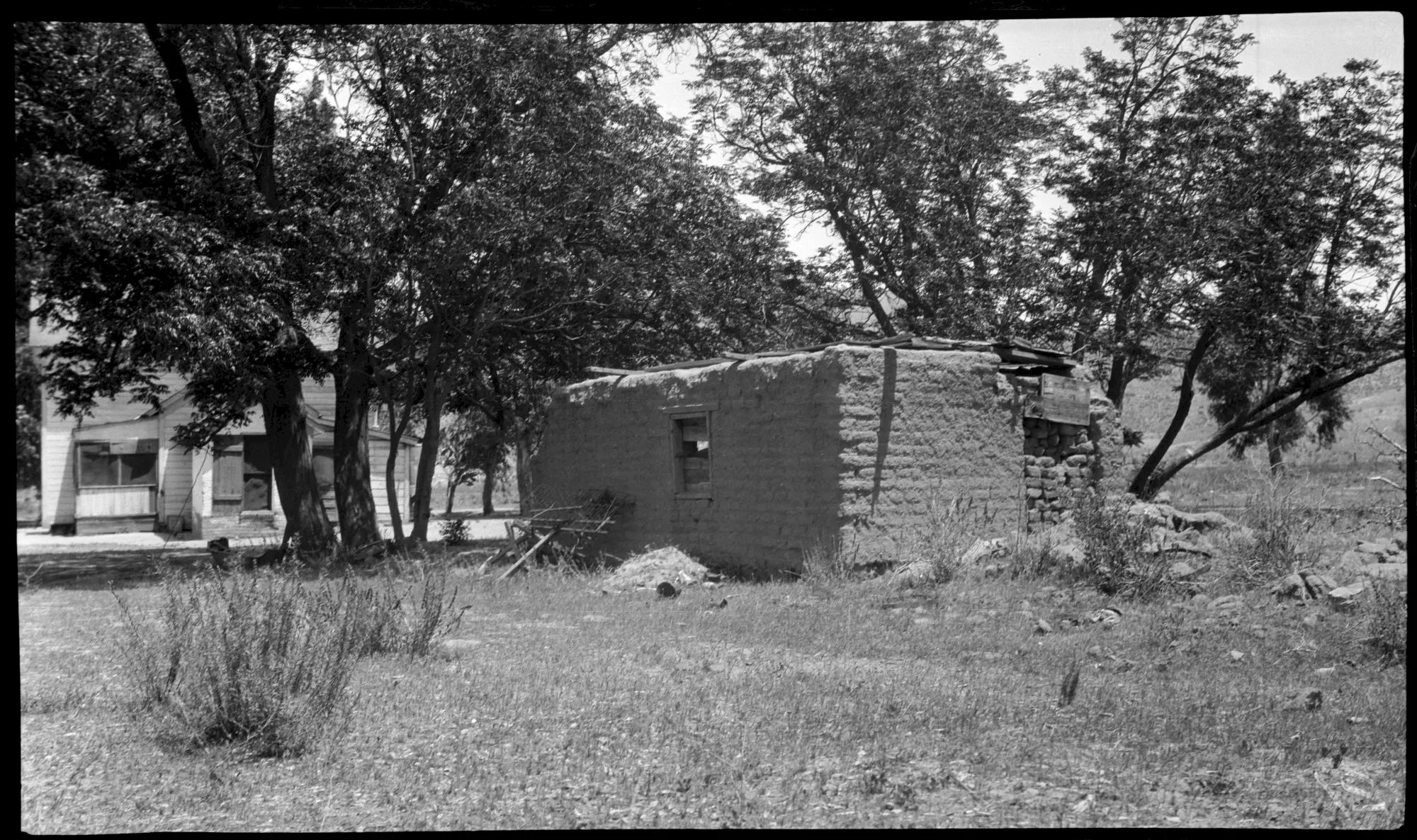 A black-and-white rural yard with a small house, a stone-and-adobe wall building, trees, and scattered tools on grass.