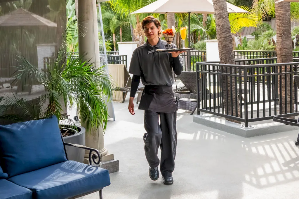 A waiter walks through a sunny tropical resort patio holding a tray with drinks and food, surrounded by palm trees and lounge chairs.