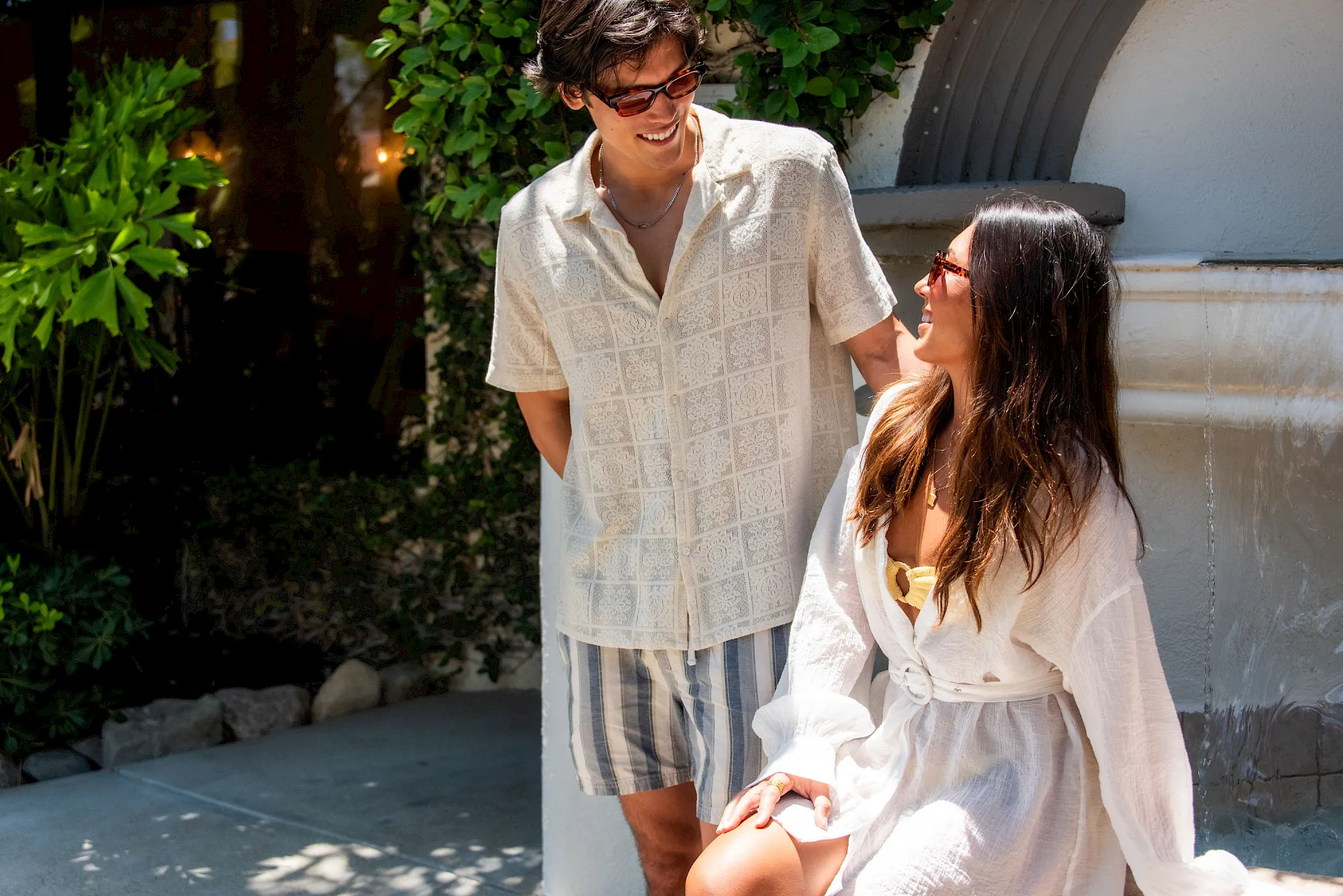 A couple wearing sunglasses in white outfits smiles together outdoors beside lush green plants, enjoying sunny patio vibes.