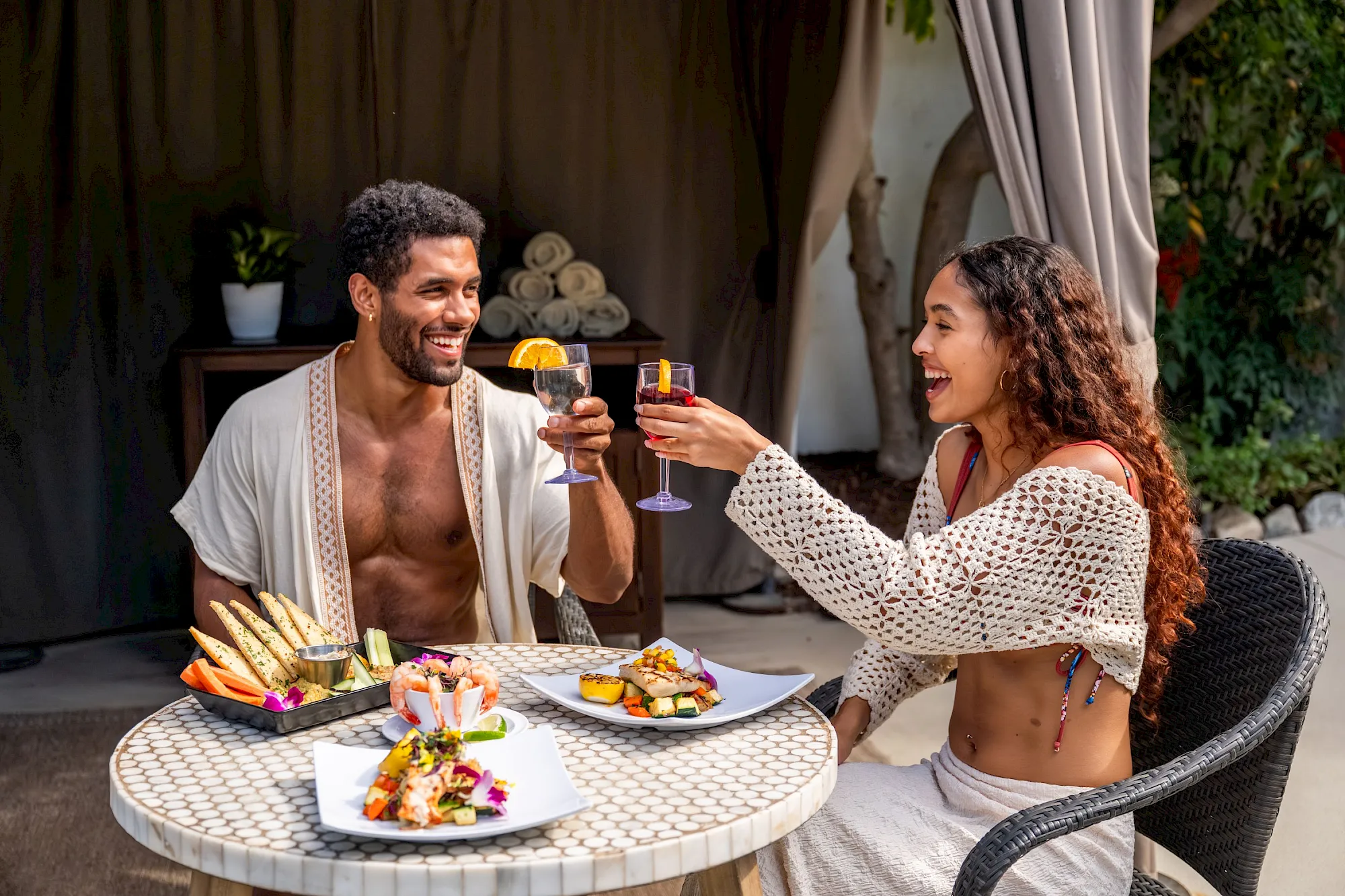 A smiling couple cheers with drinks at an outdoor table set for a romantic meal, enjoying each other&rsquo;s company and the warm evening.