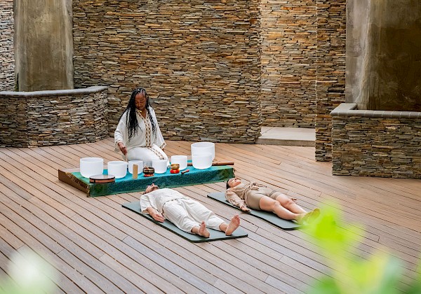 A woman leads a yoga/meditation session in a stone-walled studio, with two people lying on mats and candles on a table.