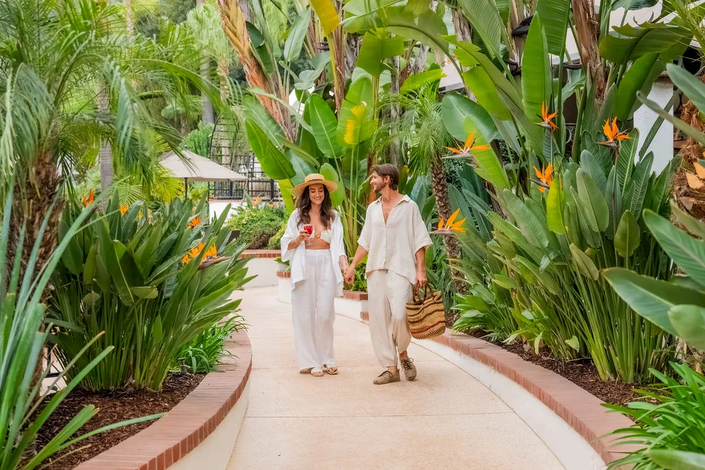 A couple walks hand in hand along a sunny garden path lined with lush tropical plants and orange bird-of-paradise flowers, smiling.