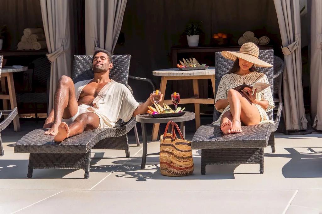 A couple lounges poolside on cushioned chairs with drinks and snacks, reading and relaxing in the sun.