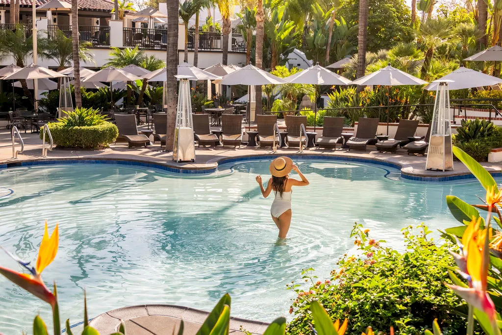 A woman splashes in a resort swimming pool, surrounded by palm trees, greenery, and sun loungers under white umbrellas.