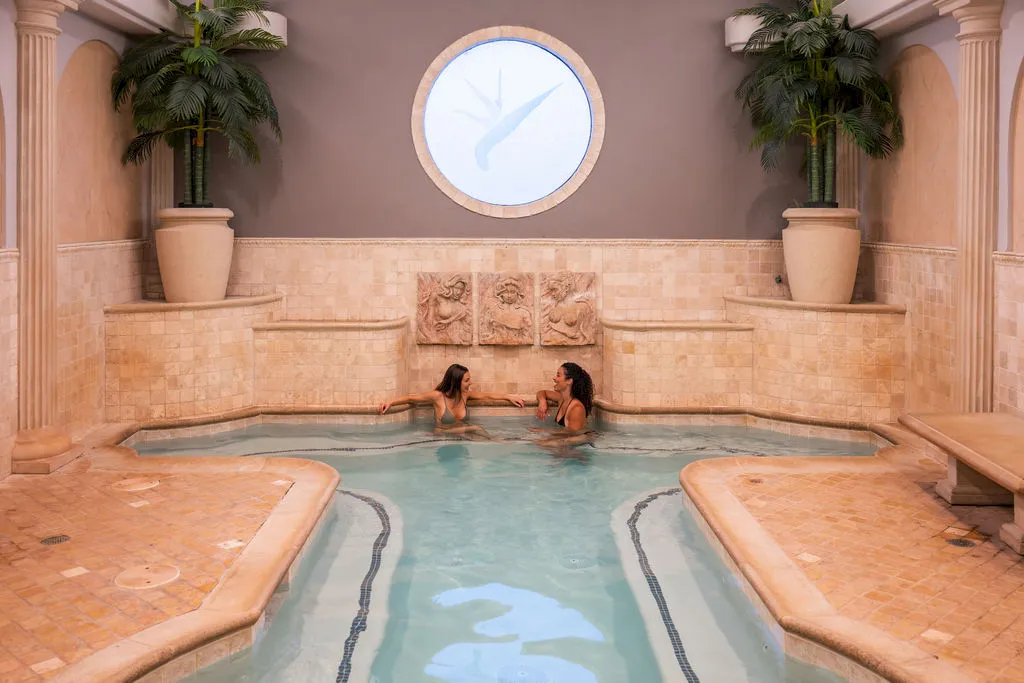 A spa pool with two women in the water, surrounded by palm trees and ornate columns in a warm indoor setting.