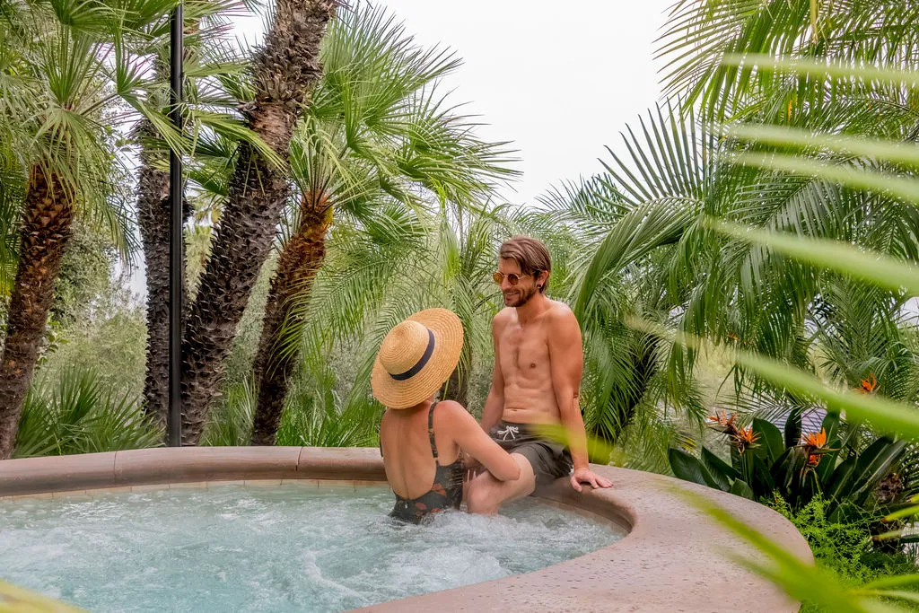 A couple relaxes in a shallow outdoor hot tub surrounded by palm trees, with a man shirtless and a woman wearing a hat.