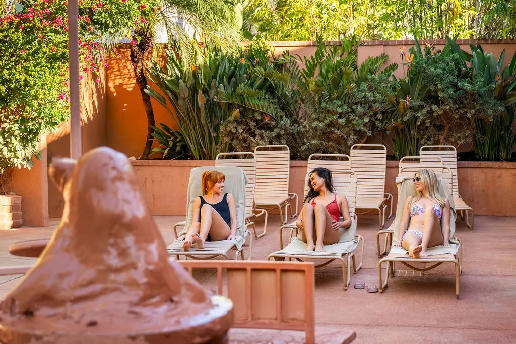 An outdoor poolside scene with four women relaxing on lounge chairs among tropical plants and sunshine, ending in a casual conversation.