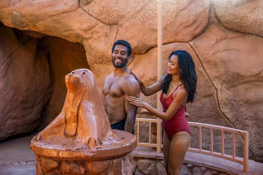 A smiling couple enjoys a warm outdoor spa, with a man and woman standing beside a steaming hot-tub basin in a rocky setting.