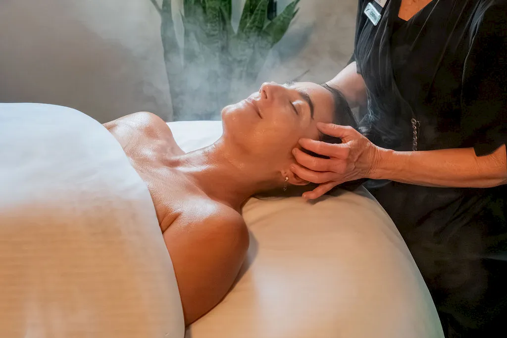 A therapist performs a massage on a shirtless man&rsquo;s face and neck on a massage table in a spa.