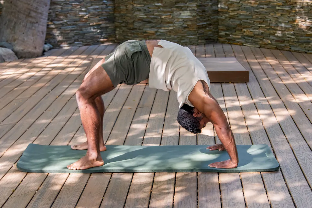 A man doing a yoga plank stretch on a mat outdoors, bending down with hands and feet planted on a wooden deck, ending in a downward-dog pose.