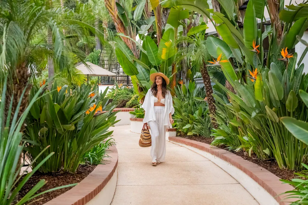A woman in a white outfit and straw hat walks along a garden path lined with lush green tropical plants and orange bird-of-paradise flowers.