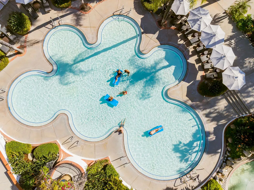 Aerial view of a hotel pool with swimmers relaxing on blue mats, surrounded by umbrellas and sunbathers, in bright sunshine.
