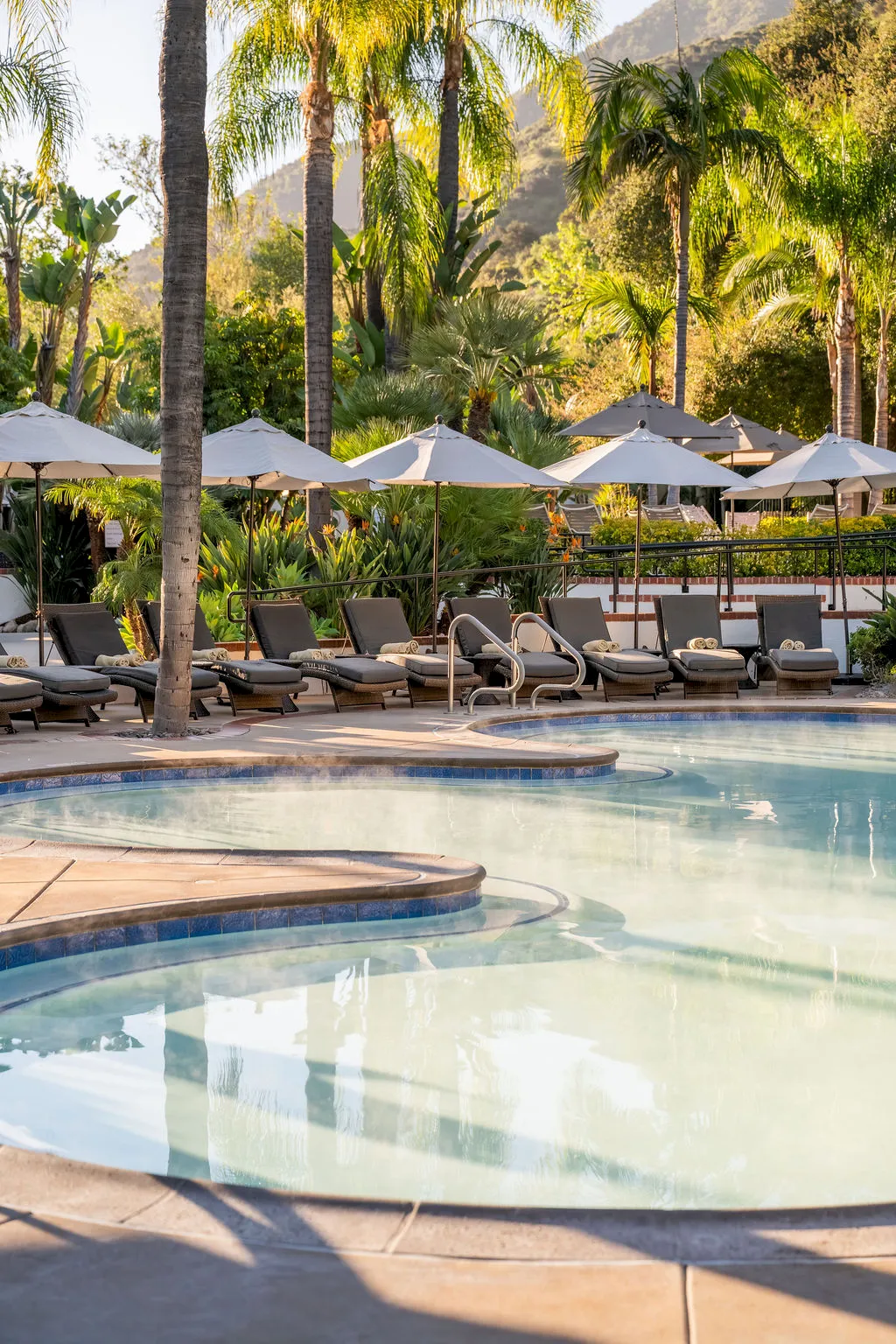 An outdoor tropical resort pool with lounge chairs, white cabanas, palm trees, and mountains in the background under warm sunlight.